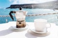 Moka pot espresso maker and cup of coffee on table with coastline and ocean in background Royalty Free Stock Photo