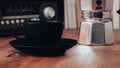 Moka pot, vintage radio, a black coffee cup and saucer on a brown wooden table, with books in the background, vintage look