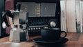 Moka pot, vintage radio, a black coffee cup and saucer on a brown wooden table, with books