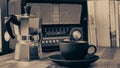Moka pot, vintage radio, a black coffee cup and saucer on a brown wooden table, with books in the background,