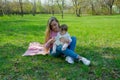 Mom with baby in bright clothes on a pink plaid on the green right. Family resting in the park on a warm day