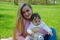 Mom with baby in bright clothes on a pink plaid on the green right. Family resting in the park on a warm day