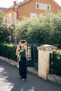 Mom with a baby on a sling walks down the street past the fence of a house with greenery