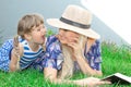 Mom blonde in a hat and her daughter are lying on the grass and reading a book, happy family. Royalty Free Stock Photo