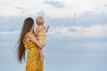 Mom in bright dress and with long hair holds baby and looks at the sea. Maternity concept. Vacation with child Royalty Free Stock Photo