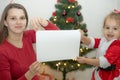 Mom and daughter dressed in red hold a white sheet of paper against the background of the Christmas tree Royalty Free Stock Photo