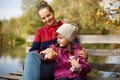 Mom and daughter sitting on wooden bench, looking at beautiful river, girls dressed casually, mother hugs her little girl and Royalty Free Stock Photo