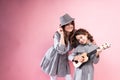Mom in a gray hat smiling stands on a pink background next to her daughter playing the ukulele
