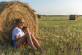 Mom and her little son are sitting in a field with hay rolls in the setting sun. A woman in denim shorts and a white t-shirt. Royalty Free Stock Photo