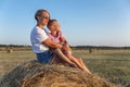 Mom and her little son are sitting in a field with hay rolls in the setting sun. A woman in denim shorts and a white t-shirt. Royalty Free Stock Photo