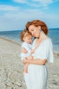 Mom is holding and smiling a little daughter 1 year old with red curly hair in light clothes in the summer on the beach.