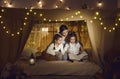 Mom and kids reading book of fairy tales sitting in blanket fort in cozy dark playroom Royalty Free Stock Photo