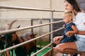 Mom with a little girl squatted in front of a corral with goats on a farm