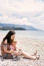 Mom puts a sandal on the little girl foot while sitting on the pebble beach