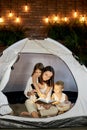 Mom reads children a bedtime story sitting in a tent at home. Mother son and daughter hug and read a book with a flashlight in Royalty Free Stock Photo