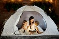 Mom reads children a bedtime story sitting in a tent at home. Mother son and daughter hug and read a book with a flashlight in Royalty Free Stock Photo