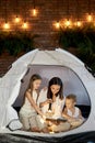 Mom reads children a bedtime story sitting in a tent at home. Mother son and daughter hug and read a book with a flashlight Royalty Free Stock Photo