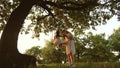 Mom shakes a healthy little daughter on swing under tree in sun. mother plays with child they are swinging on a rope on Royalty Free Stock Photo