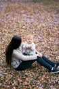 Mom sits on leaves in the park, bowing her head, holding a smiling child on her knees