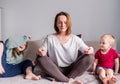 Mom sits on the sofa meditating lotus position, little children scream playing around focused calm mom meditating balancing while