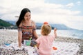 Mom smiles at the little girl with a toy mold on her head while sitting on a pebble beach