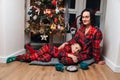 Mother and son in red pajamas sit near the Christmas tree with tea and chocolate cookies. Front view