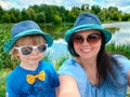 Mom and son in sunglasses in blue T-shirts in blue hats with yellow bow tie smiling on river bank. Mother and son are Royalty Free Stock Photo