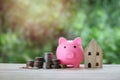 Money coin stack, pink piggy bank and wooden house over blurred green garden background, saving money and business financial