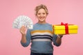 Money and gift. Portrait of beautiful happy woman with curly hair in warm sweater holding present box and dollar bills Royalty Free Stock Photo