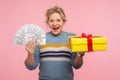 Money and gift. Portrait of shocked excited woman with curly hair in warm sweater holding present box and dollar bills Royalty Free Stock Photo