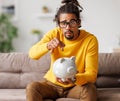 money problems. Poor upset african american man putting coin in ceramic piggy bank