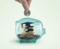 Money savings for education concept. Hand putting a coin into a green piggy bank with books, diploma and graduation cap toy inside Royalty Free Stock Photo