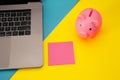 Moneybox near laptop and pink sticky note, place for text. Finance and budget concept. Piggy bank in blue color with