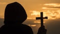 A monk in a hood with a crucifix in his hands stands against the backdrop of a dramatic sky at sunset