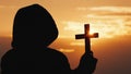 A monk in a hood with a crucifix in his hands stands against the backdrop of a dramatic sky at sunset