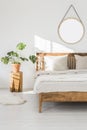 Monstera plant on a tree trunk night stand and a round mirror on a white wall in a sunlit bedroom interior with wooden furniture