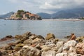 A woman is sitting on the stone and reading a book near the water on the background of the island of St. Stephen on a sunny summer