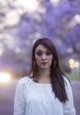 Moody image of beautiful woman in white dress standing in street surrounded by purple Jacaranda trees Royalty Free Stock Photo