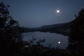 Moonlight from a glowing fullmoon reflected in a lake at midnight with mountains and trees around Royalty Free Stock Photo