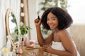 Morning beauty routine. Attractive black lady applying mascara, sitting at dressing table, looking in mirror at home Royalty Free Stock Photo