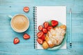 Morning coffee cup with fresh croissant, strawberry and empty notebook on turquoise rustic table from above, tasty breakfast