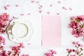 Morning coffee mug for breakfast, empty notebook and pink flowers on white wooden table, top view, flat lay style. Woman working