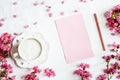 Morning coffee mug for breakfast, empty notebook and pink flowers on white wooden table, top view, flat lay style. Woman working