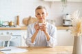 Attractive mature woman starting day with coffee or tea while sitting at wooden table in kitchen Royalty Free Stock Photo