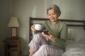 Morning lifestyle portrait of attractive and happy middle aged woman on her 50s having coffee on bed using internet mobile phone Royalty Free Stock Photo