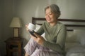 Morning lifestyle portrait of attractive and happy middle aged woman on her 50s having coffee on bed using internet mobile phone Royalty Free Stock Photo