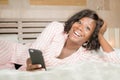 Morning lifestyle portrait of young beautiful and happy afro American woman in pajamas on bed using internet mobile phone in the Royalty Free Stock Photo