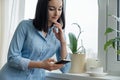 Morning portrait of young smiling woman in shirt at home near the window with cup of coffee reading text on smartphone Royalty Free Stock Photo