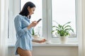 Morning portrait of young smiling woman in shirt at home near the window with smartphone reading text, on the windowsill cup of Royalty Free Stock Photo