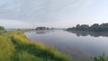 Morning on the river. Light fog and soft sunshine. Tall grass and trees grow along the banks and are reflected in the calm water.